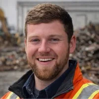 A smiling person with a beard, wearing a dark shirt and an orange high-visibility safety vest at a construction site.