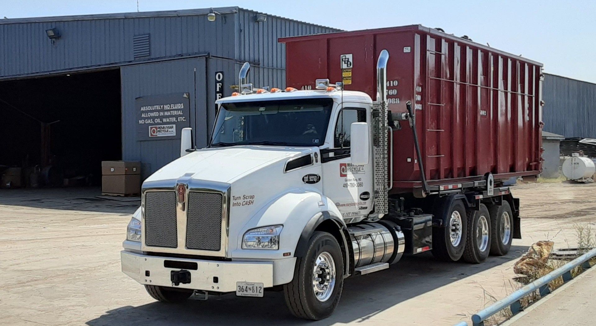 A white Kenworth truck with a large, maroon roll-off dumpster container parked in an industrial yard.
