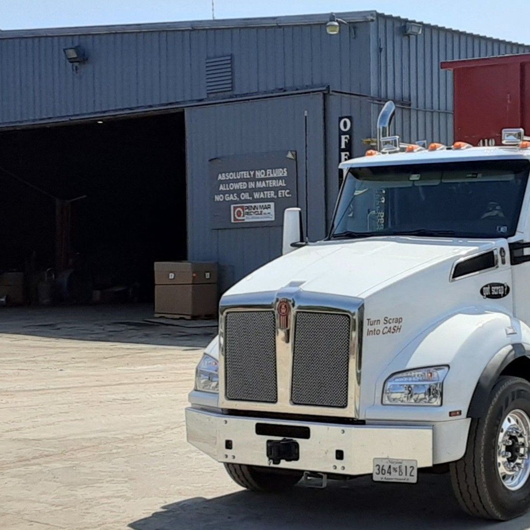A white semi-truck parked in front of a gray metal industrial building with a loading bay.