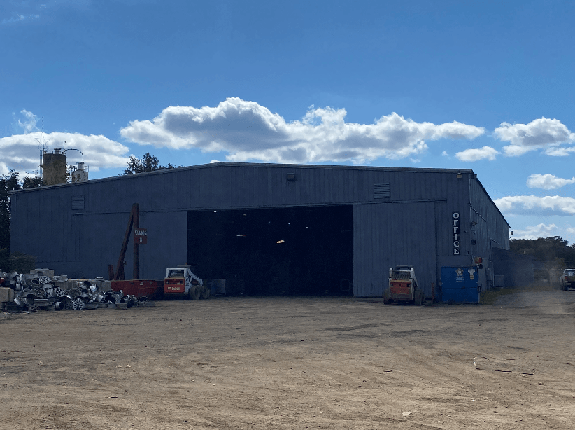 A large grey warehouse building with a wide open bay door, parked construction equipment, and a blue office container.