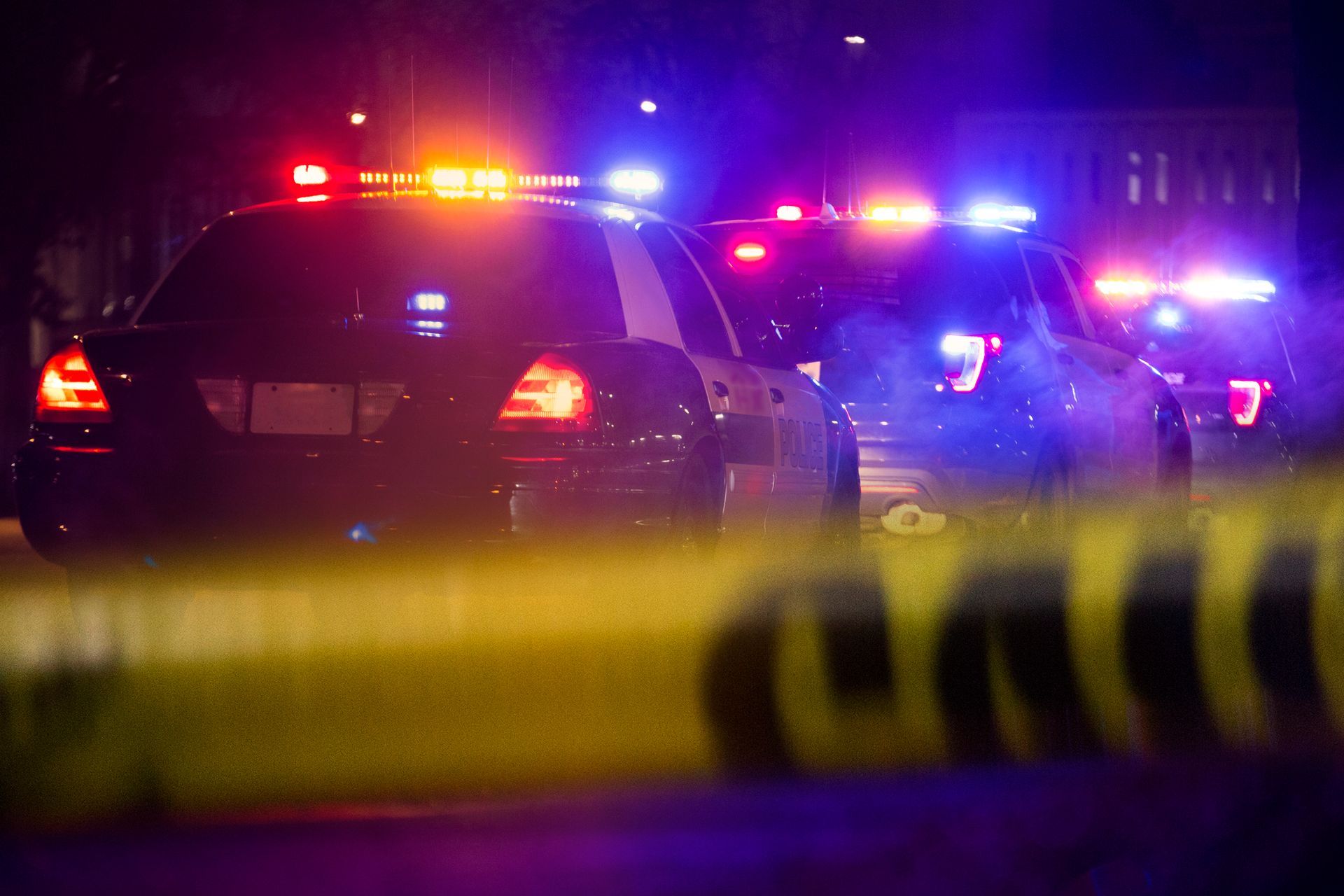 Police cars with flashing red and blue lights behind yellow caution tape at night