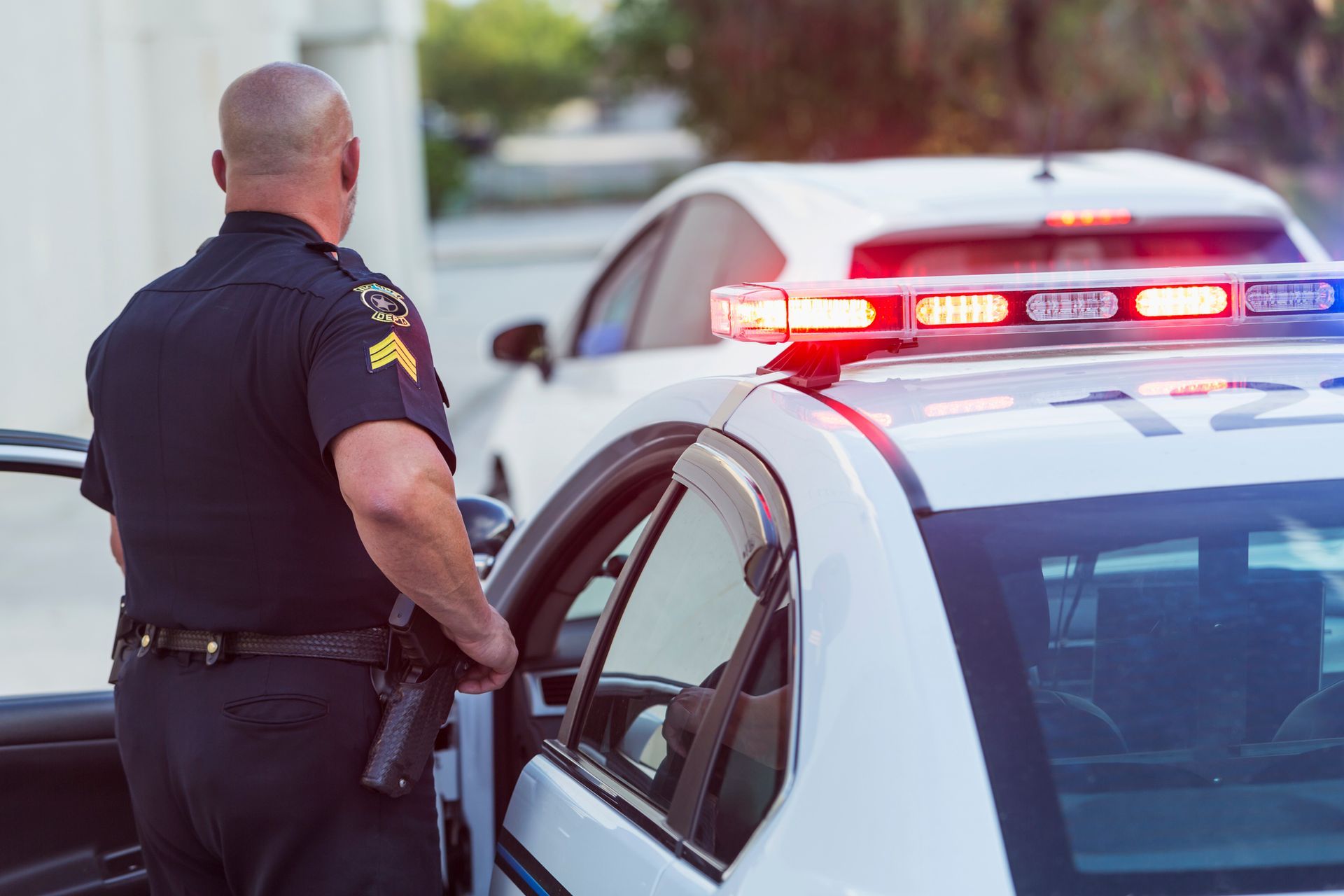Police officer stands beside a patrol car with flashing red and blue lights during a traffic stop.