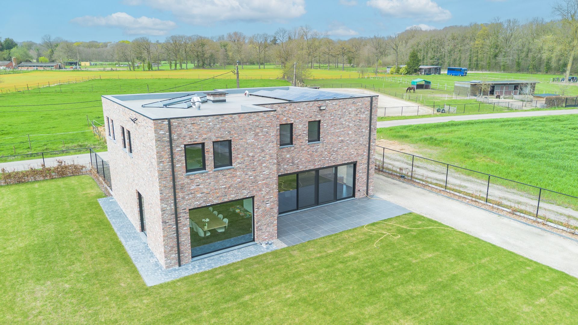 Moderne, twee verdiepingen tellende bakstenen woning met grote ramen, omgeven door een groen gazon in een landelijke omgeving.