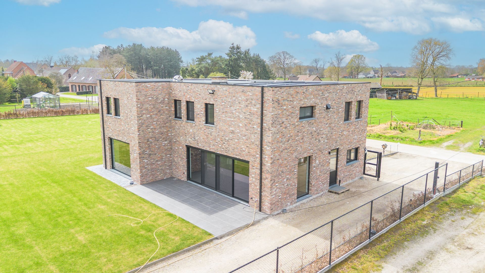 Moderne, twee verdiepingen tellende bakstenen woning met grote ramen, gelegen naast een omheinde oprit in een landelijke, groene omgeving.