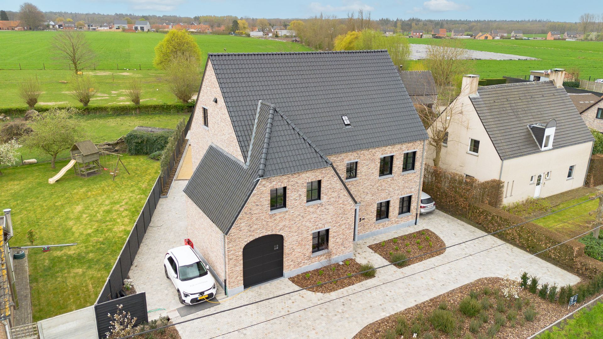 Luchtfoto van een modern bakstenen huis met een donker dak, een geparkeerde auto en groene velden in de buurt.
