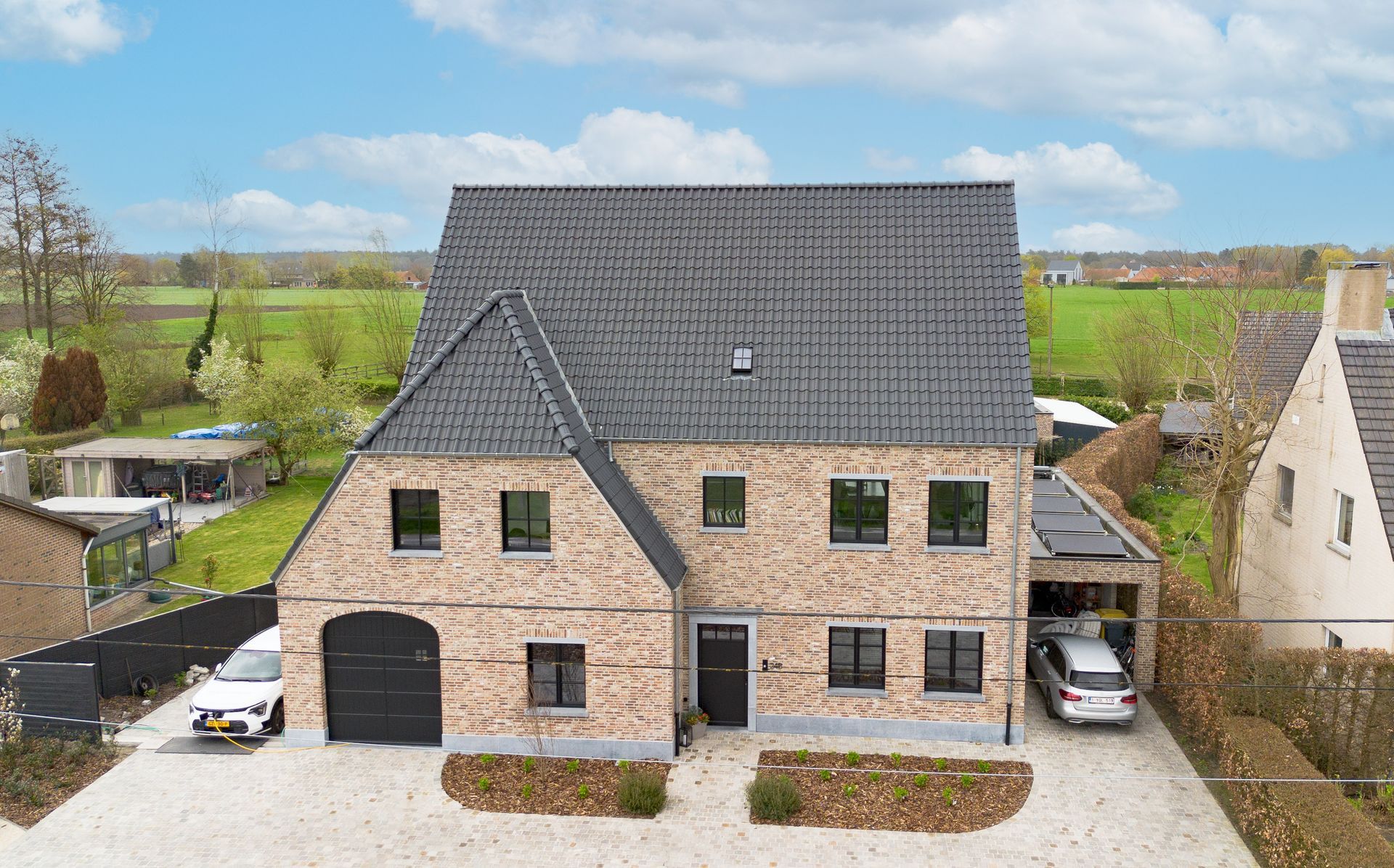 Luchtfoto van een groot bakstenen huis met een donker dak en groene luiken, omgeven door velden.
