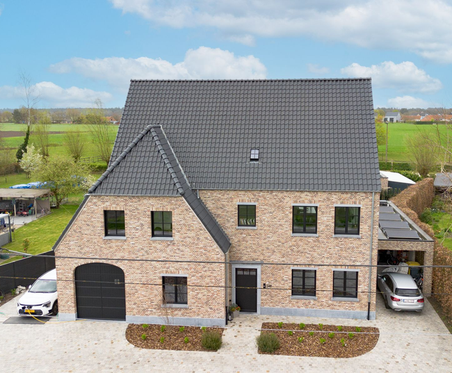 Groot bakstenen huis met donker dak, zwarte garagedeur en grindoprit in een landelijke omgeving.
