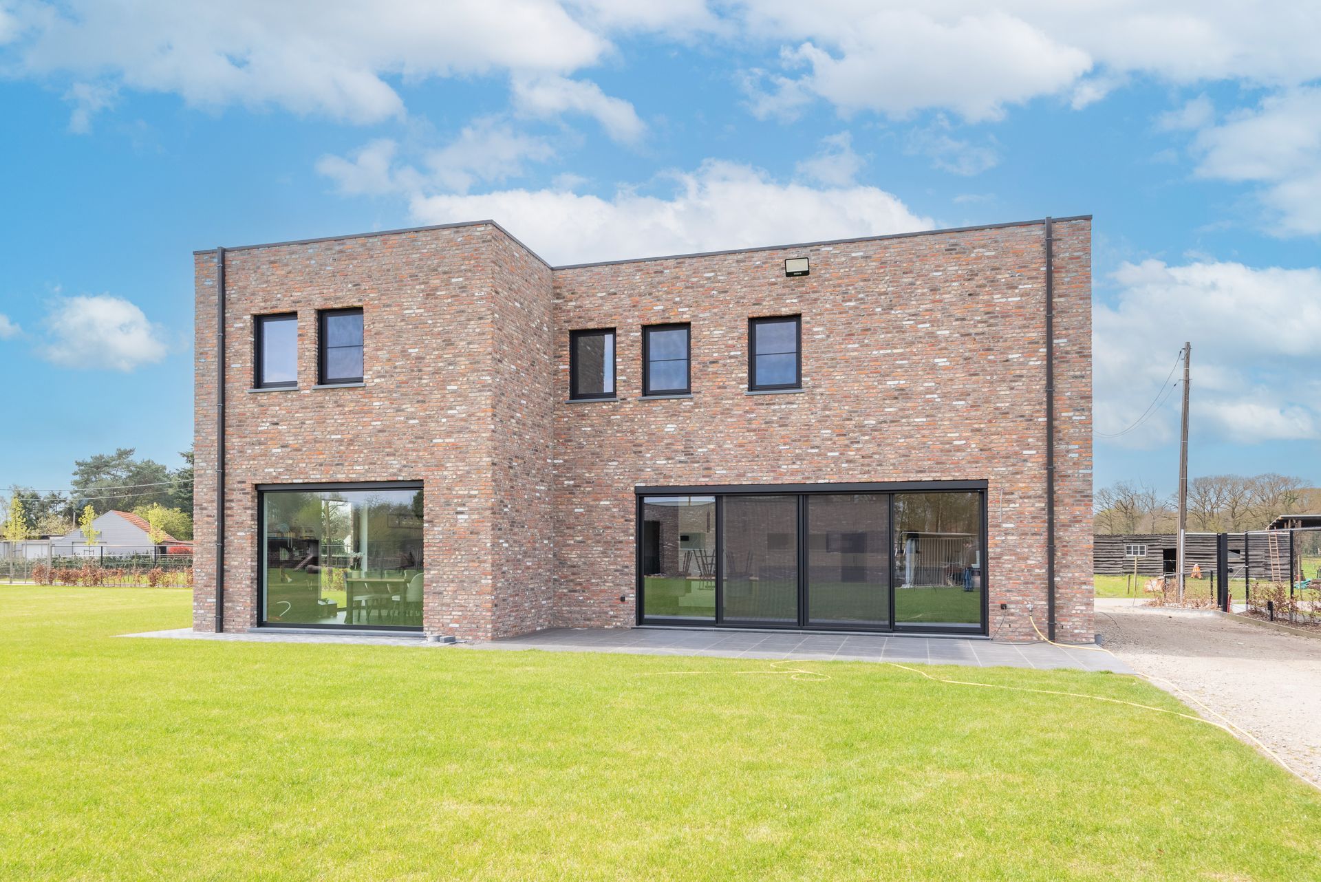 Moderne, twee verdiepingen tellende bakstenen woning met grote ramen en een groen gazon onder een blauwe hemel.