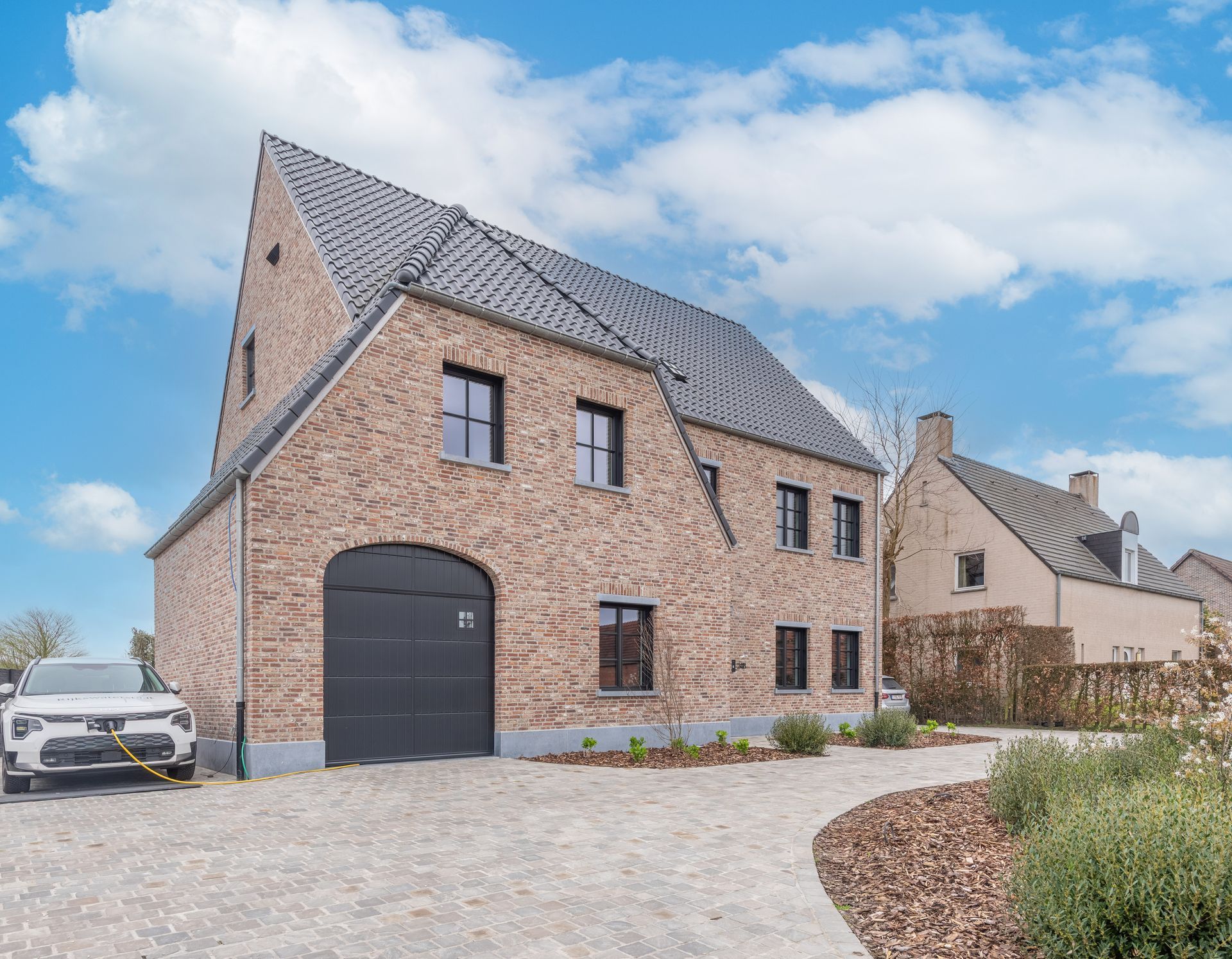 Modern bakstenen huis met grijs dak, zwarte garagedeur, grindoprit en een geparkeerde witte auto onder een bewolkte hemel.