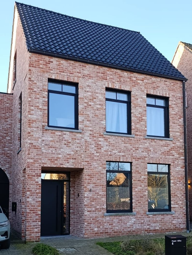 Moderne, twee verdiepingen tellende bakstenen woning met zwart dak en grote ramen onder een heldere hemel.