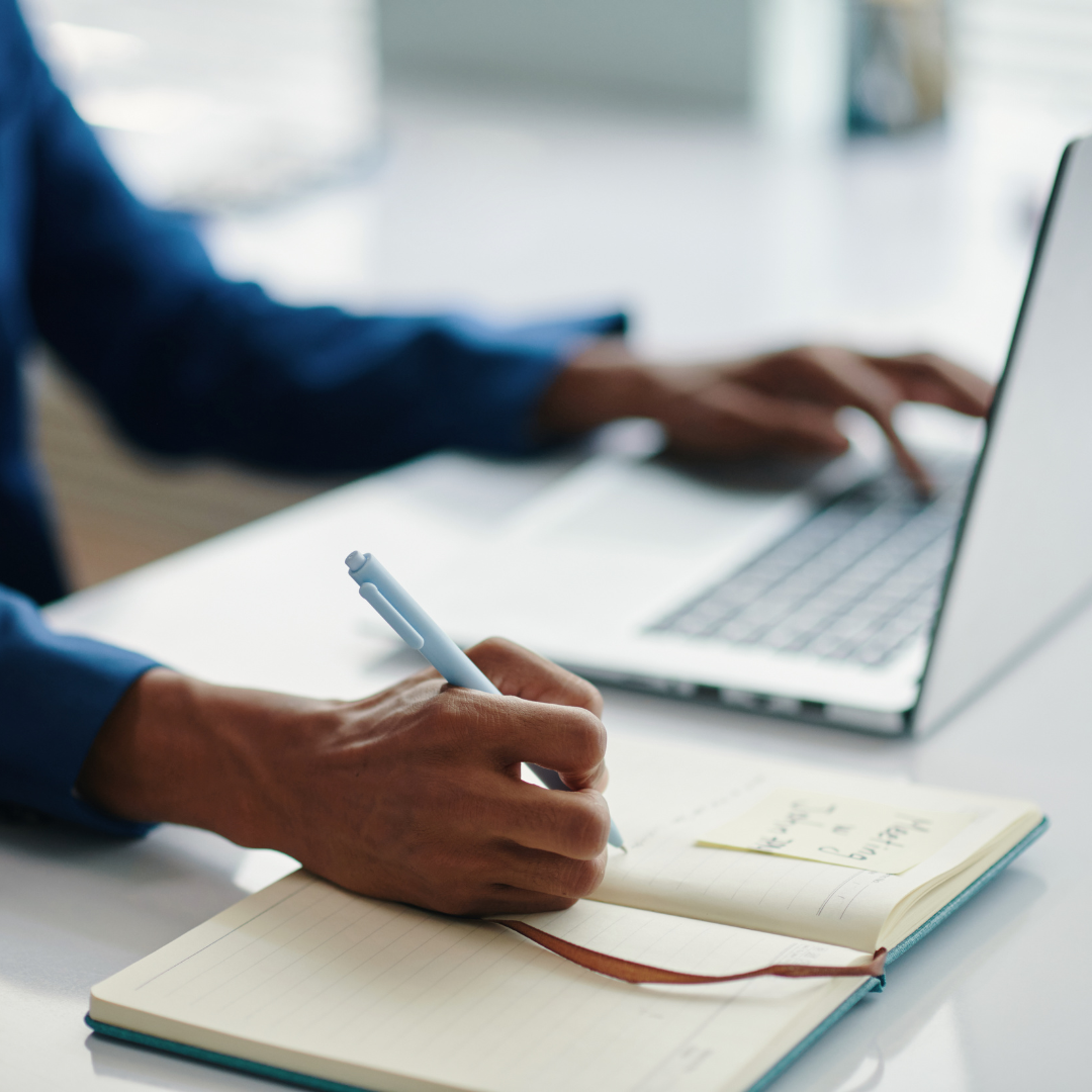 Person writing in notebook and typing on laptop at a desk with a sticky note.