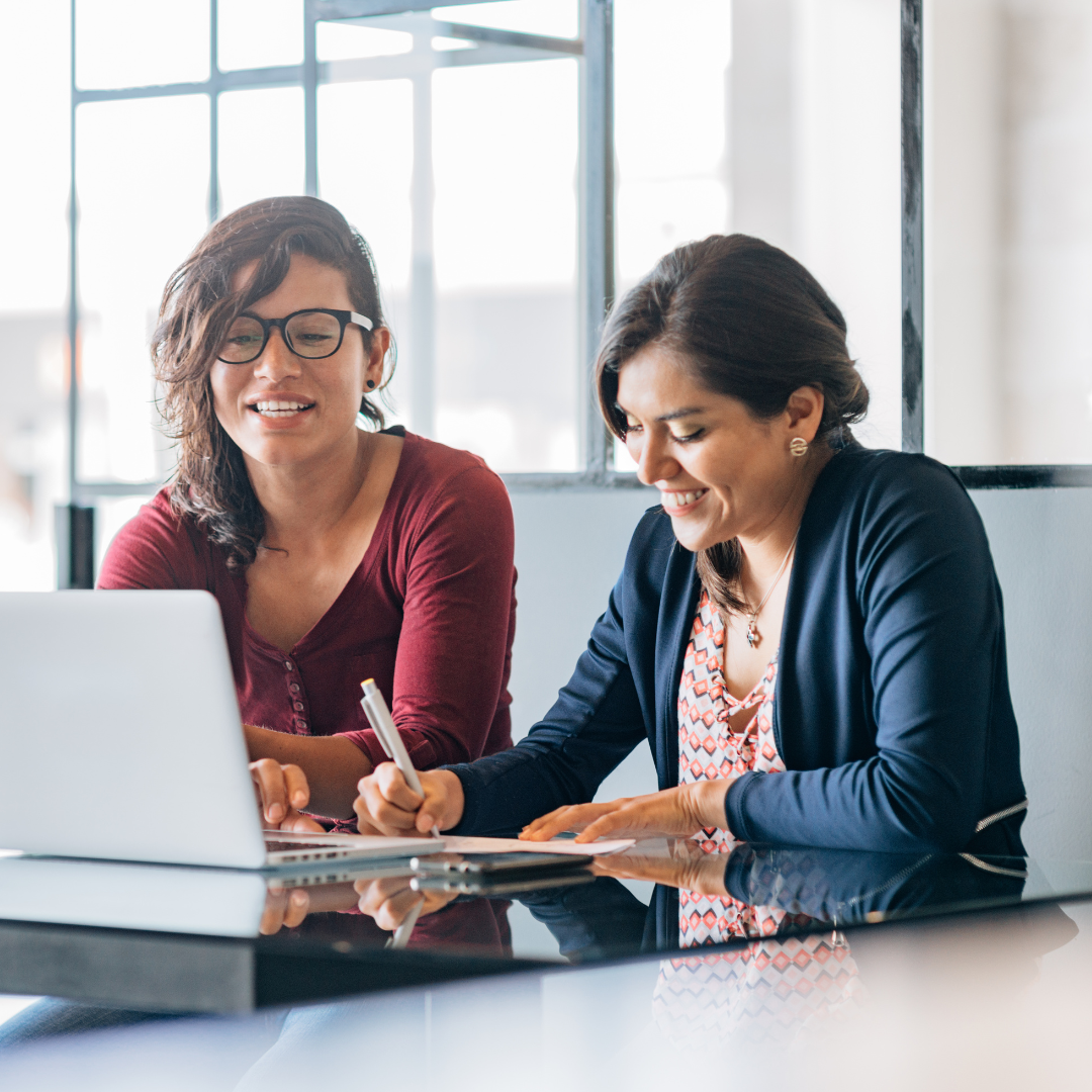 Two women smiling, working together at a table. One uses a laptop; the other writes on paper.