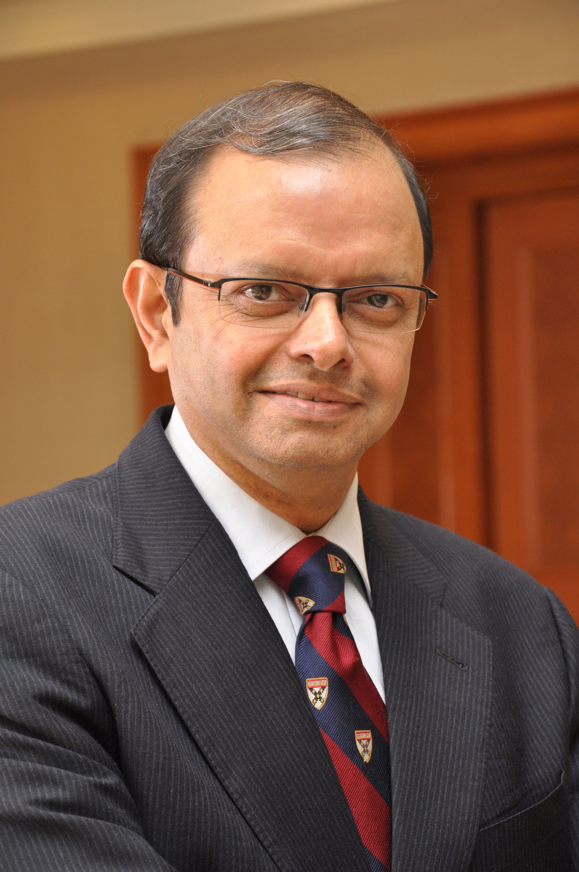 Man in a dark suit and patterned tie, wearing glasses, posed against a warm wood-toned background
