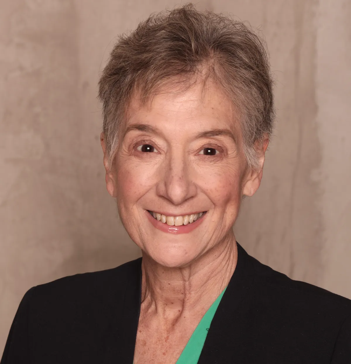 Woman with short gray hair, smiling, wearing a black blazer over a green top, indoors.