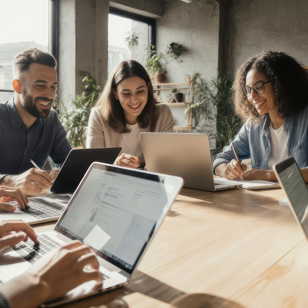 People working on laptops at a table, smiling. Bright office setting, natural light.