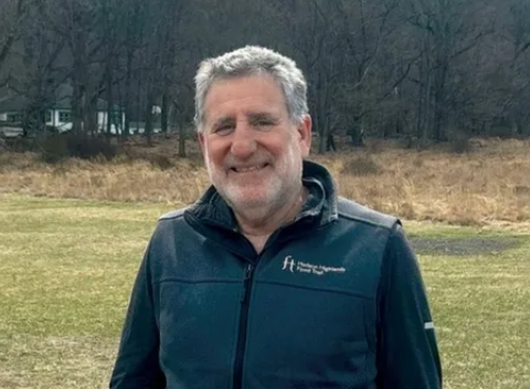 Smiling man in a navy jacket standing in a grassy field with bare trees behind him