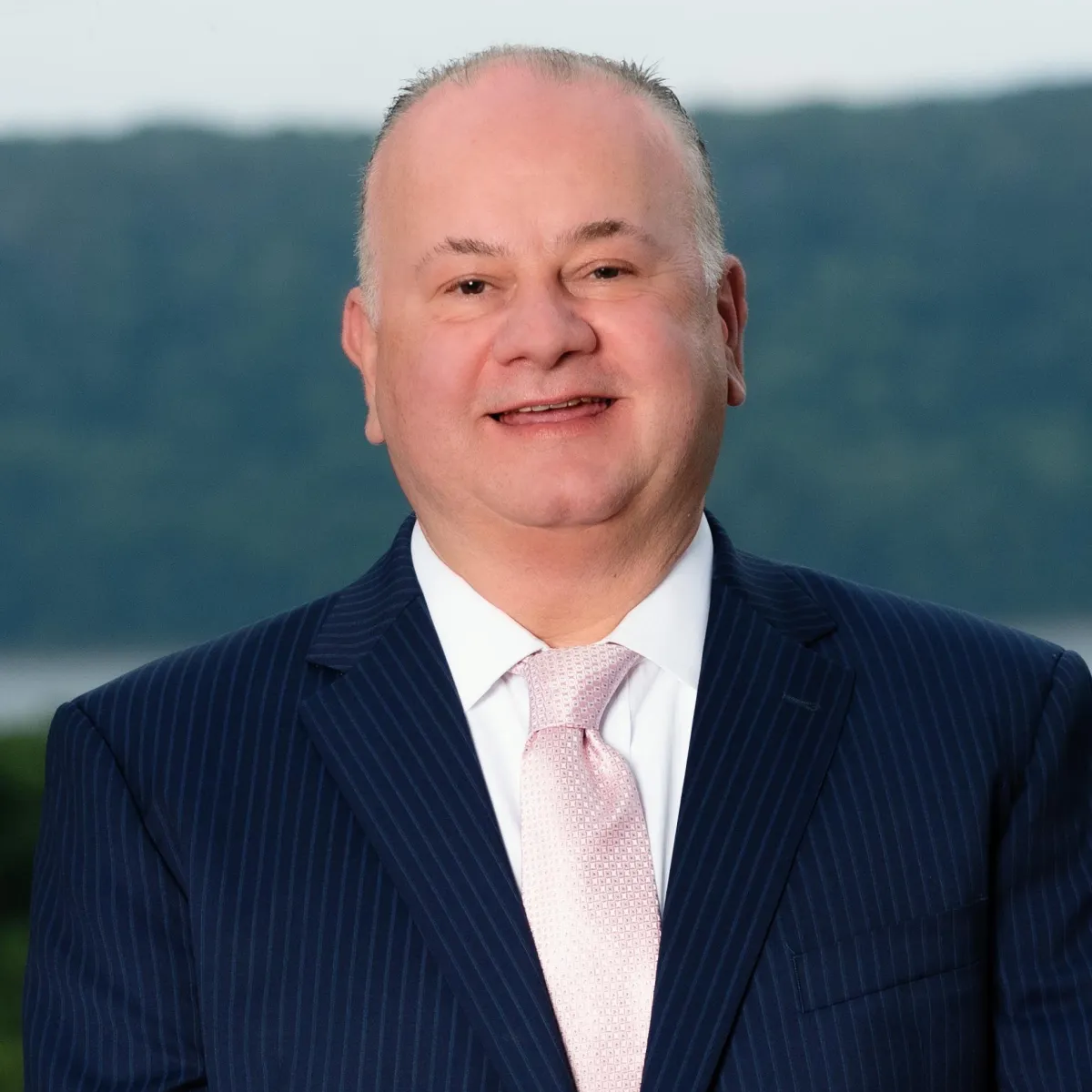 Man in navy pinstripe suit, pink tie, smiling outdoors, blurred background.