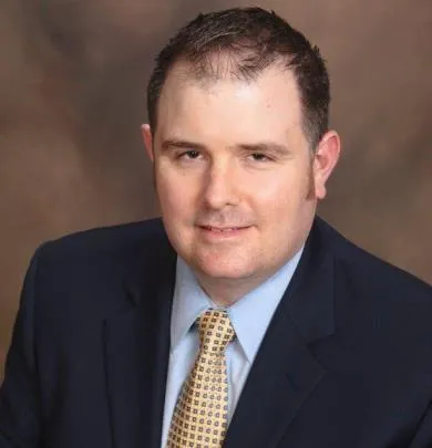 Man in navy suit, pale blue shirt, and gold tie, smiling.