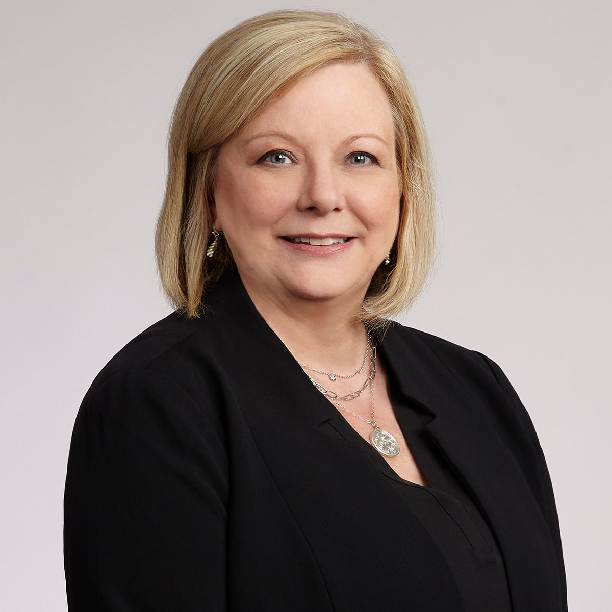 Woman with blonde hair, smiling, wearing a black blazer and necklace. Studio shot against a gray background.