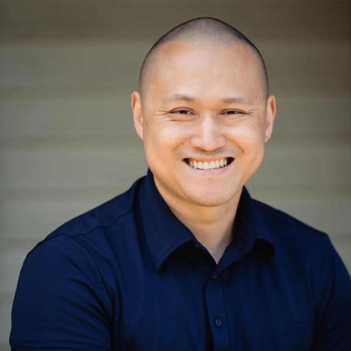 Smiling man in a dark blue shirt, bald head, outdoors against a neutral background.