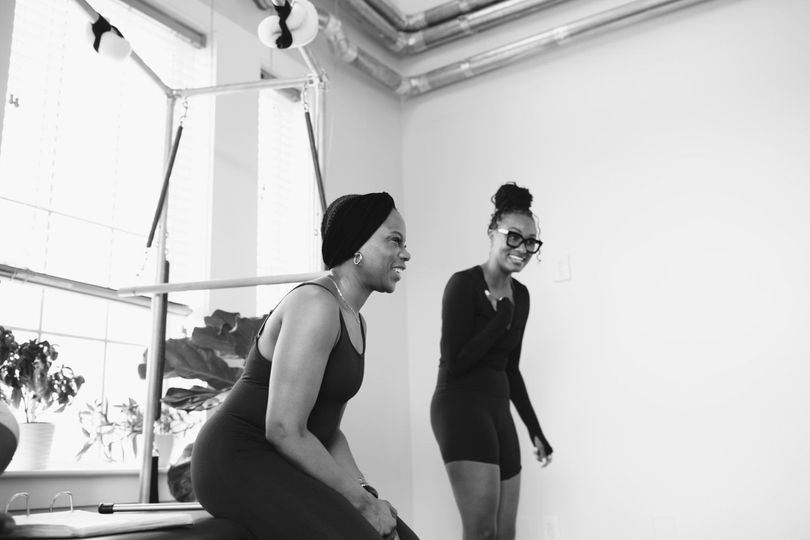 Two women in black workout clothes smiling in a bright studio by a ballet barre.