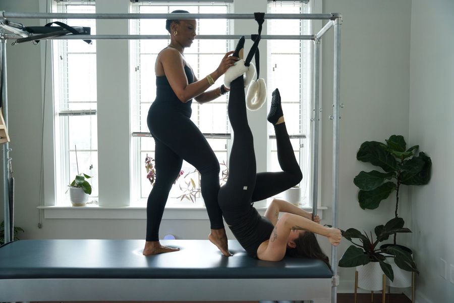 Two women practicing Pilates cadillac in a bright studio, one assisting the other in straps.
