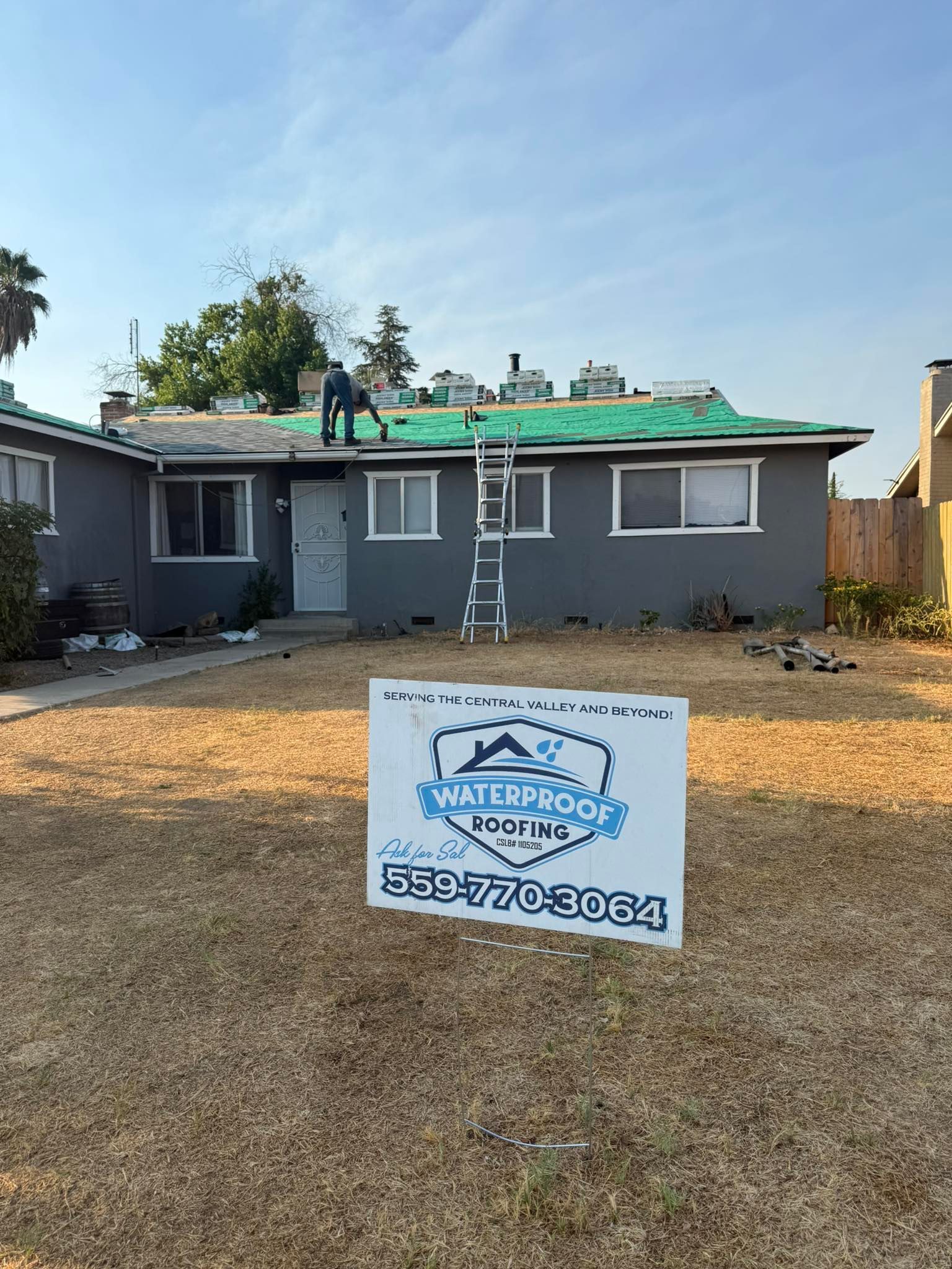 A worker repairs a gray house roof with green underlayment, with a Waterproof Roofing sign on the dry lawn in the foreground.