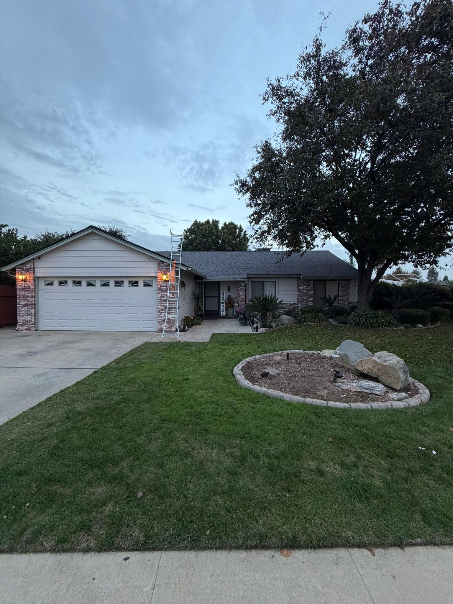 A single-story, light-colored suburban house with a two-car garage, a green lawn, and a large tree at dusk.