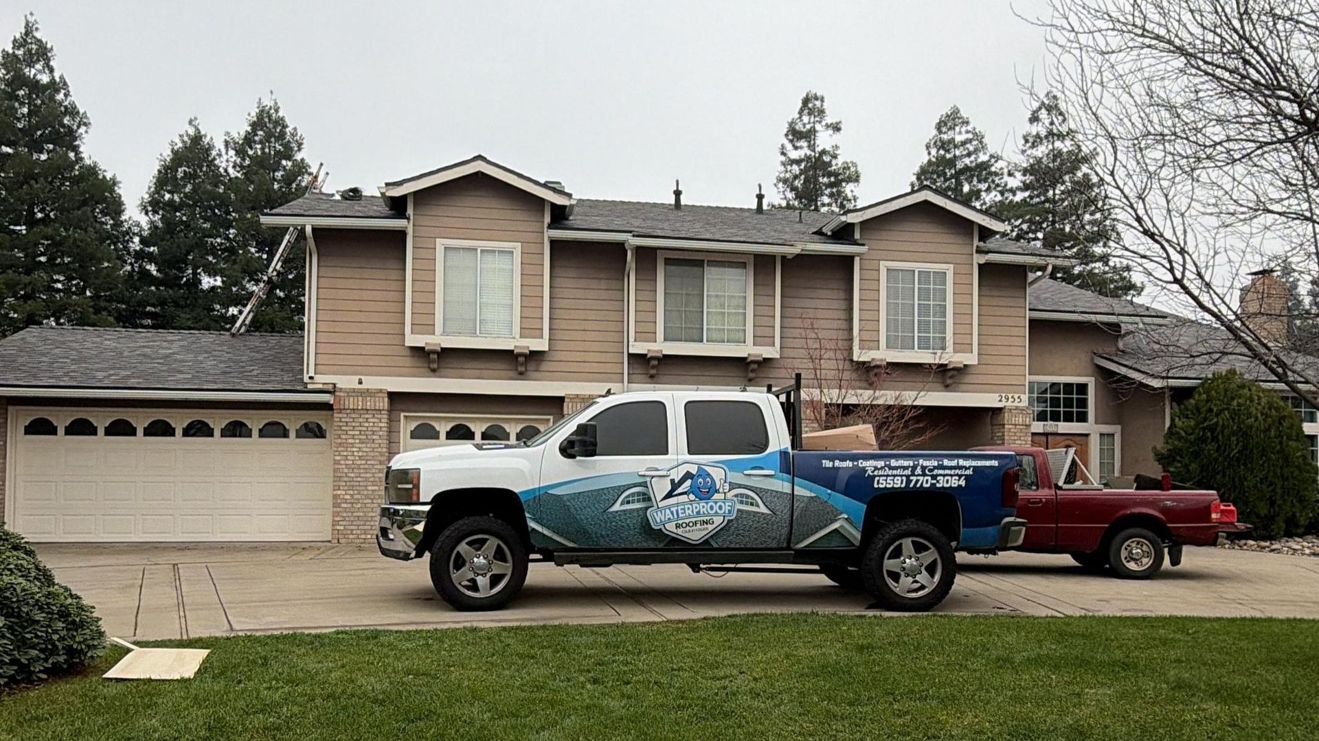 A white truck with blue and grey graphics parked in front of a two-story tan house on a residential street.