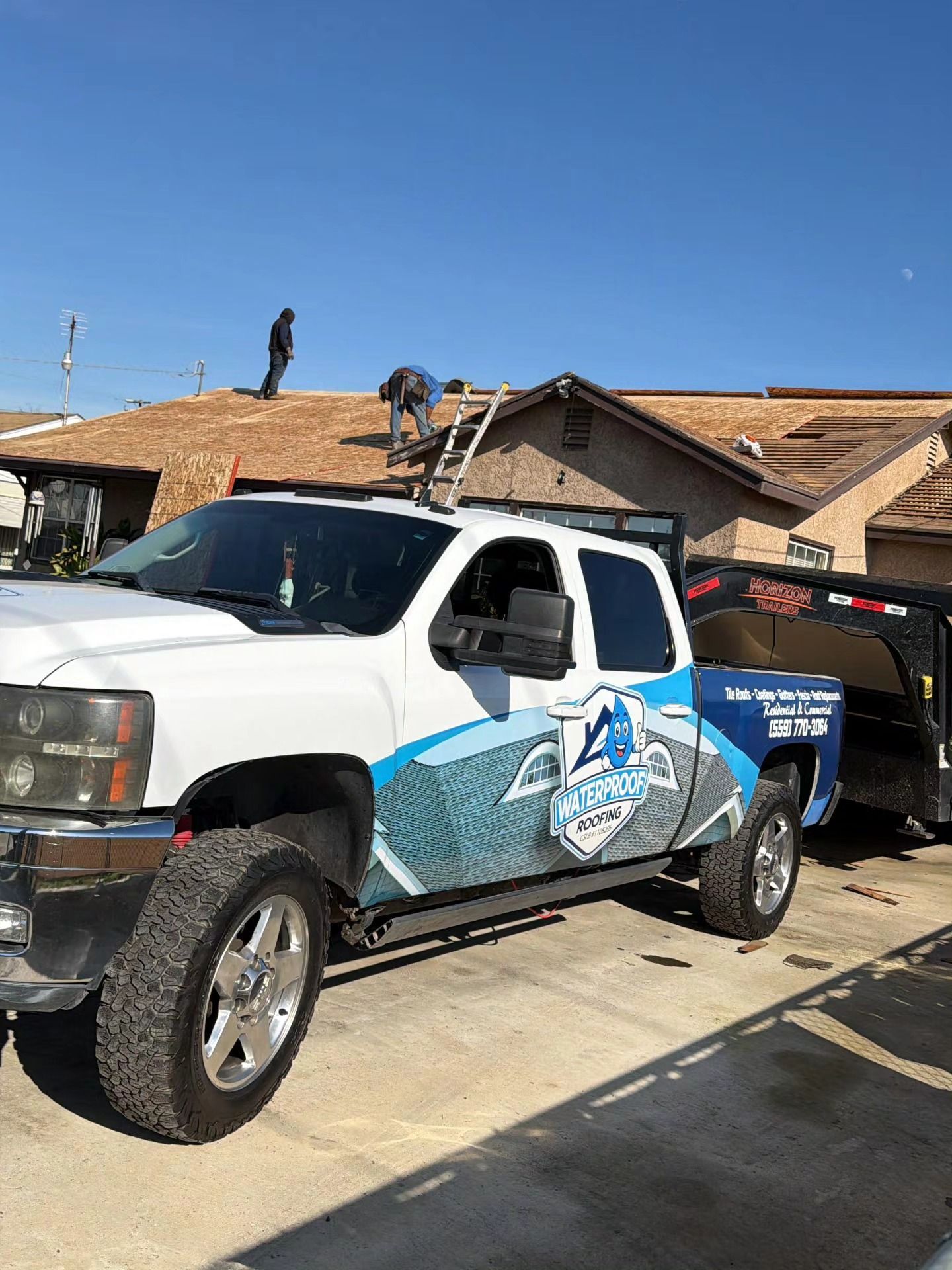 A white pickup truck with blue branded decals is parked in front of a house under construction with roofers working.