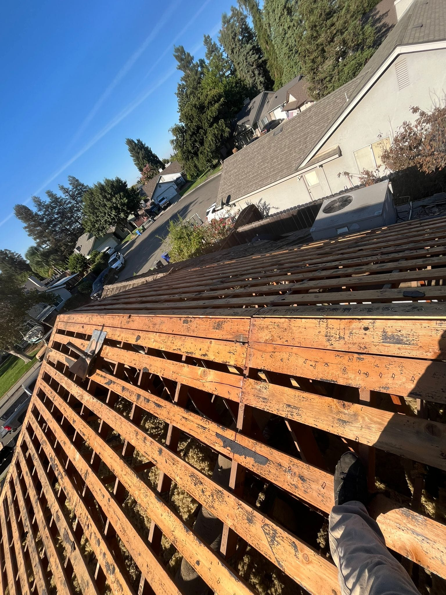 A view from a roof showing wooden slats, partially stripped roofing, and a neighborhood in the background.