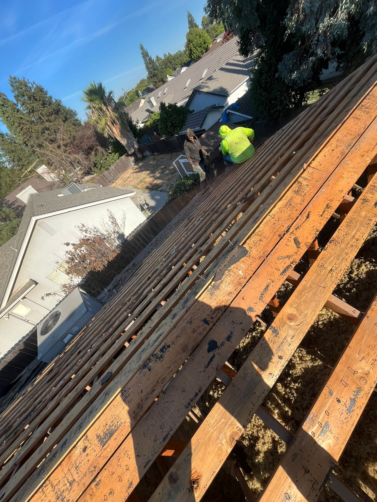 A construction worker in a high-visibility yellow vest works on a bare wood roof frame overlooking a residential area.