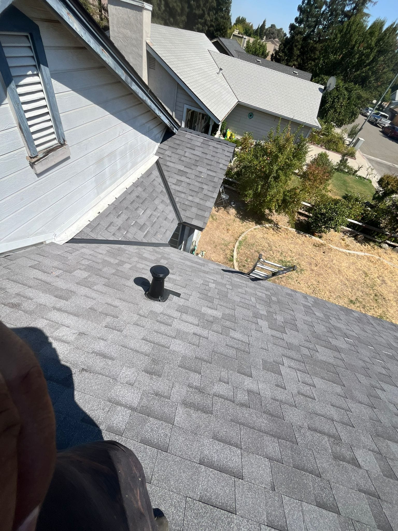 A view looking down a gray shingled roof towards a valley, with a small vent pipe and a nearby home in the background.