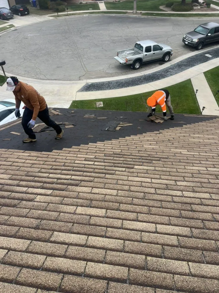 Two people in work clothing remove shingles from a residential roof while parked vehicles sit on the street below.