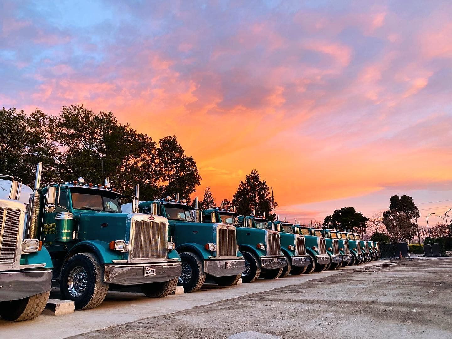 Row of green semi trucks parked along a road at sunset under a pink-orange sky Row of green semi trucks parked along a road at sunset under a pink-orange sky