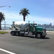 Green cement mixer truck parked on a sunny road by palm trees with a bridge and city skyline in the background
