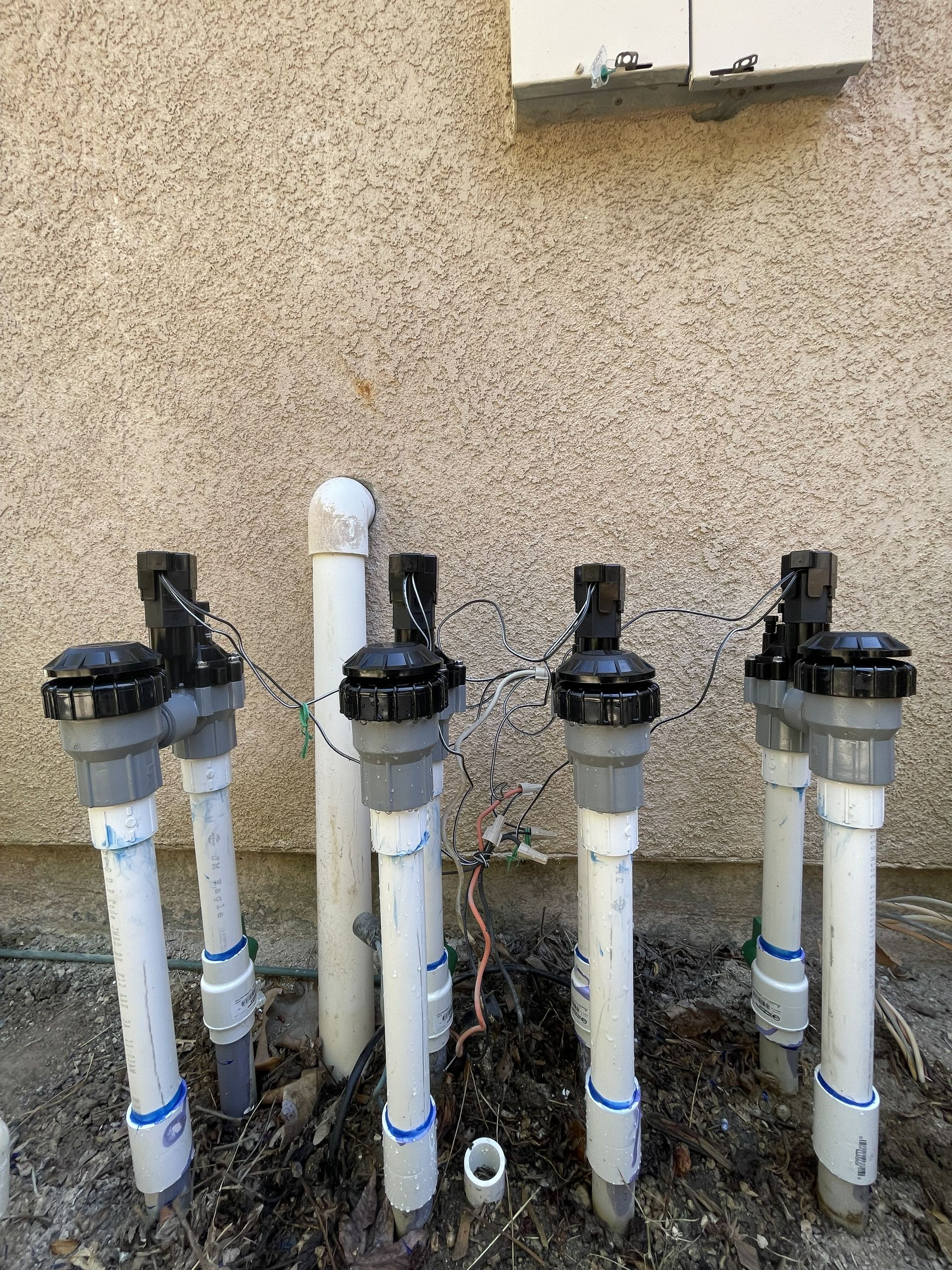 Four white PVC pipes with black valves against a beige stucco wall outdoors.