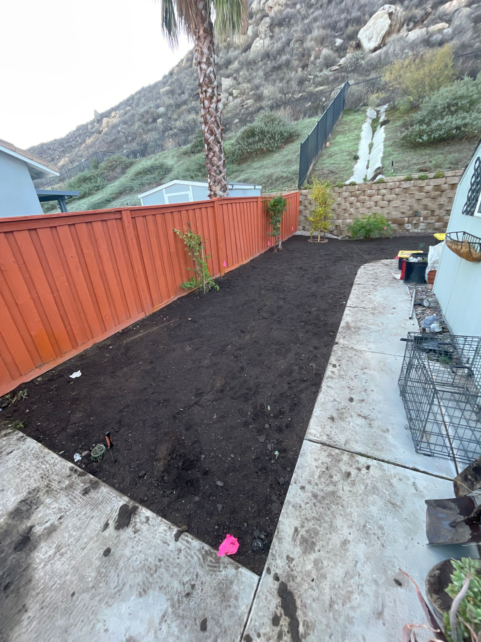 Small backyard garden bed with fresh dark soil beside an orange fence and concrete patio, mountains in background