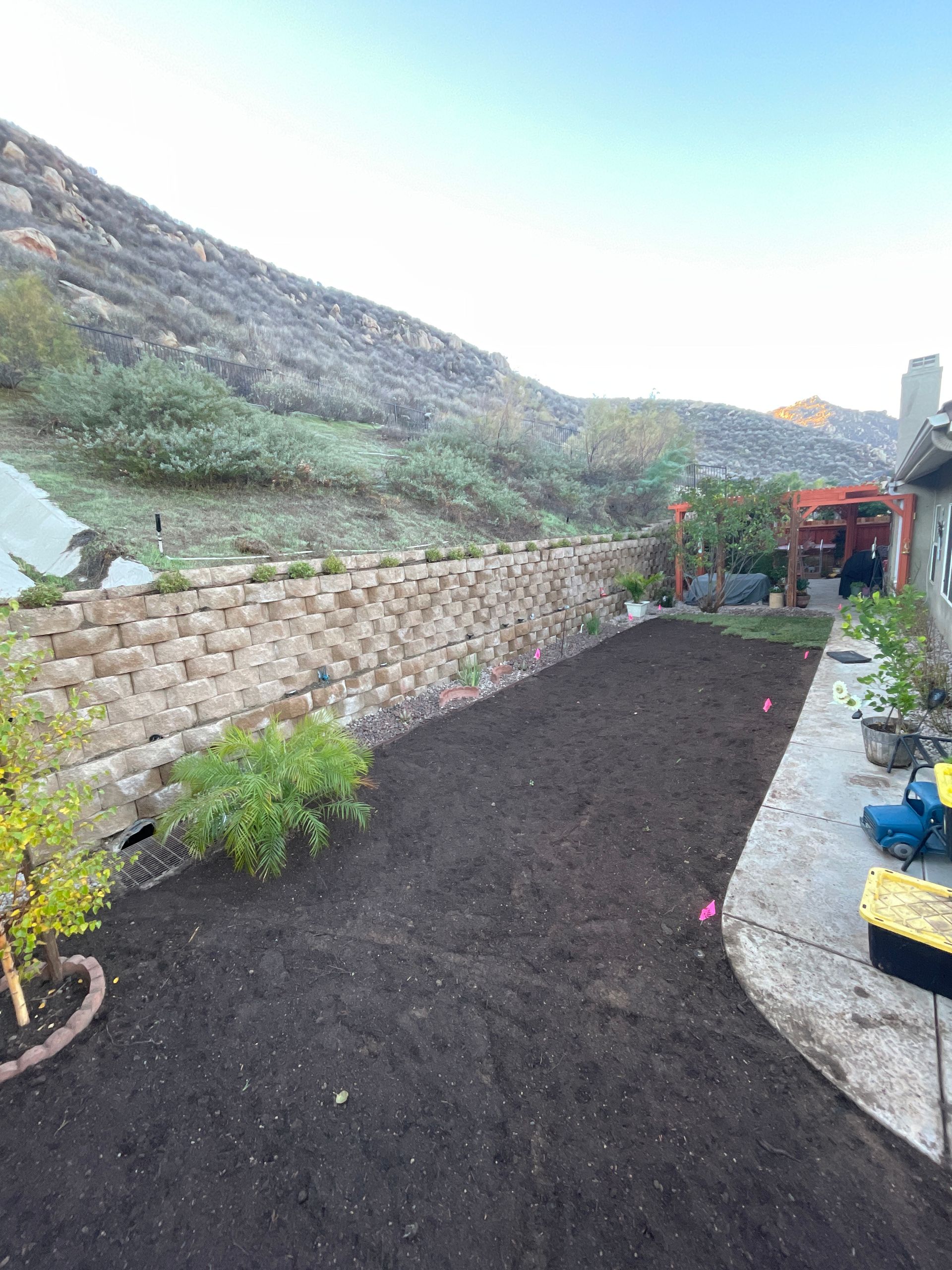 Small landscaped garden with dark soil beds, stone walls, potted plants, and a hillside backdrop.