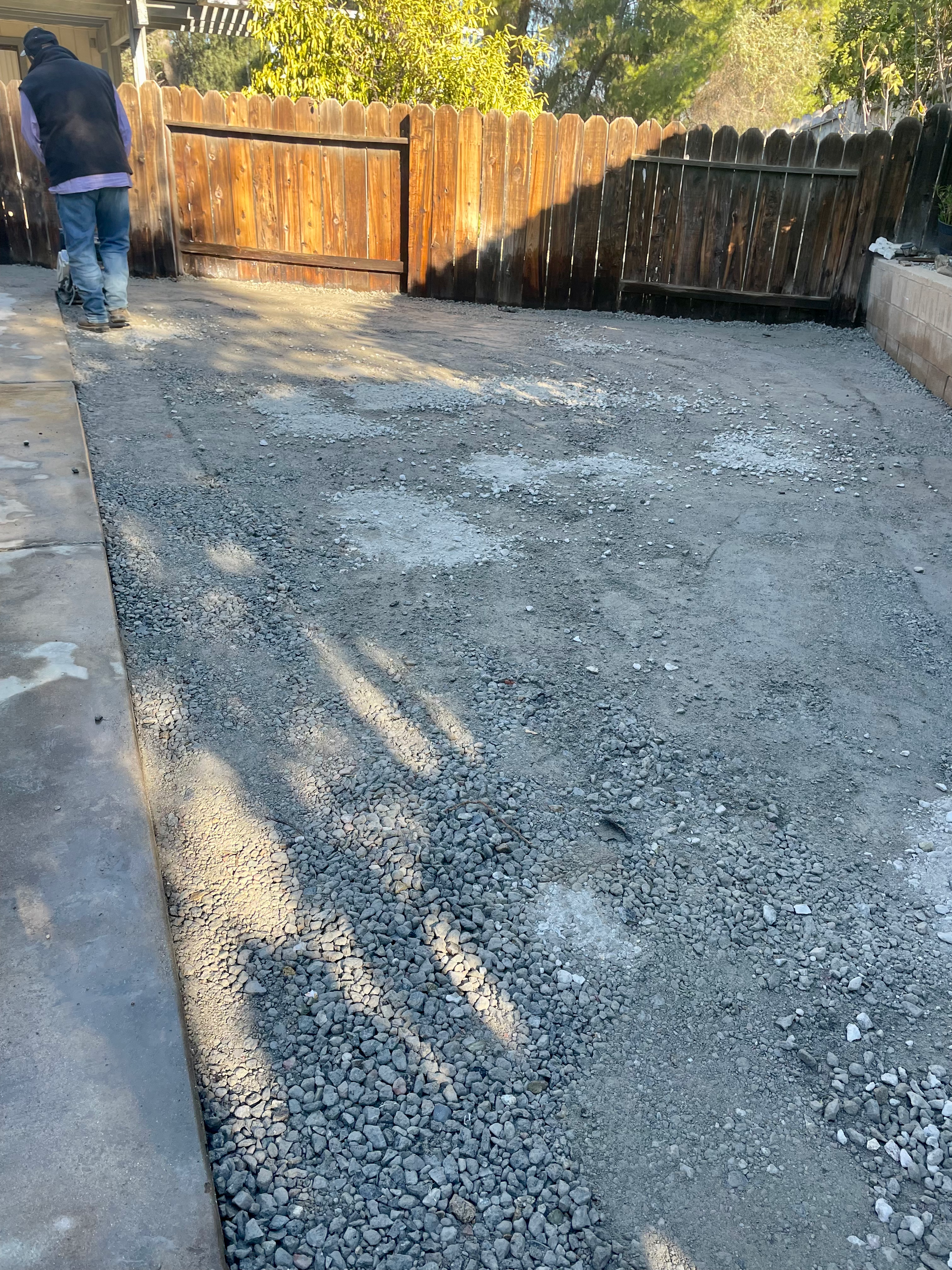 Gravel-covered backyard beside a concrete patio and wooden fence, with one person standing at left.