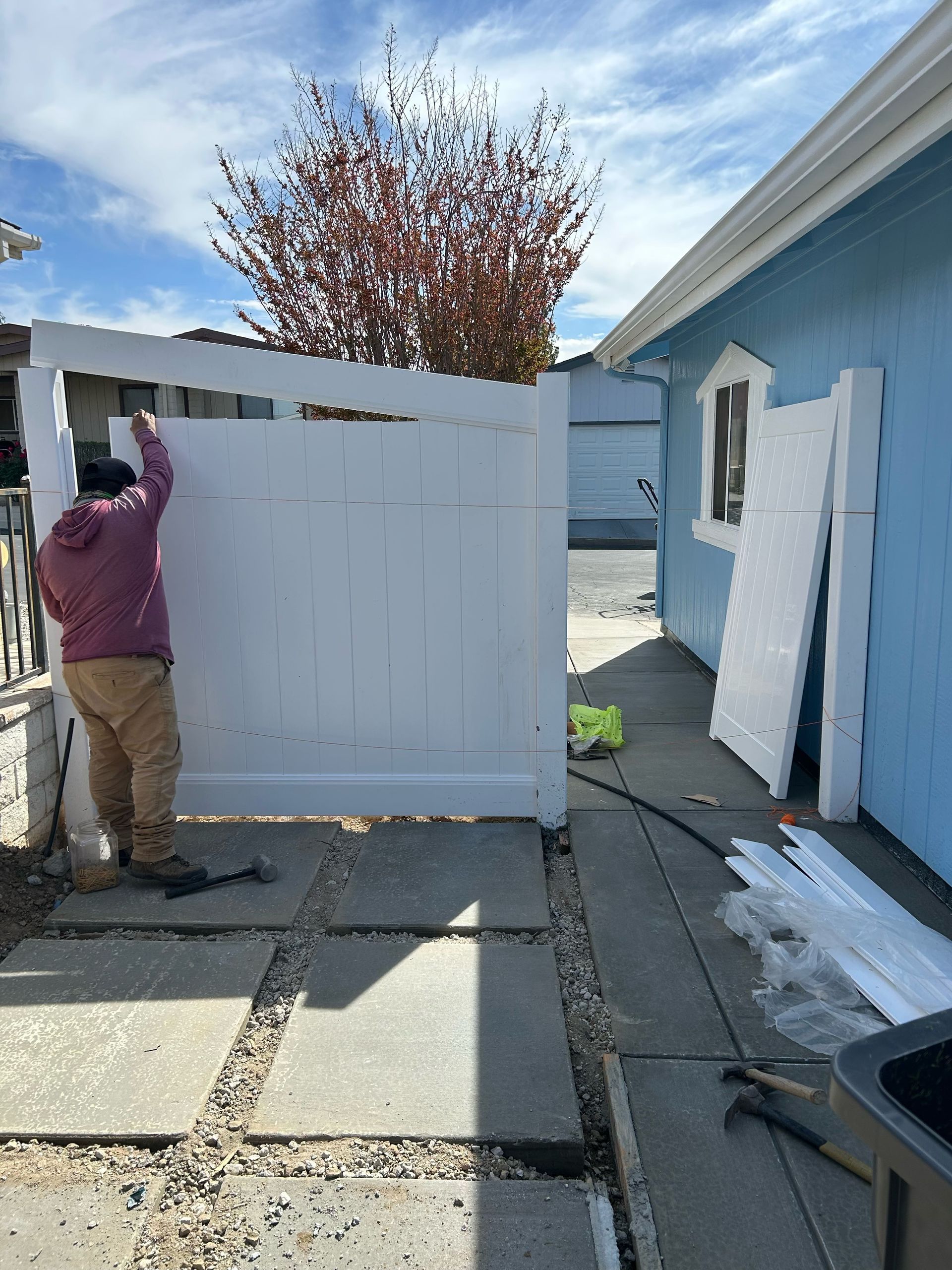 Person installing a white fence gate beside a blue house on a sunny day