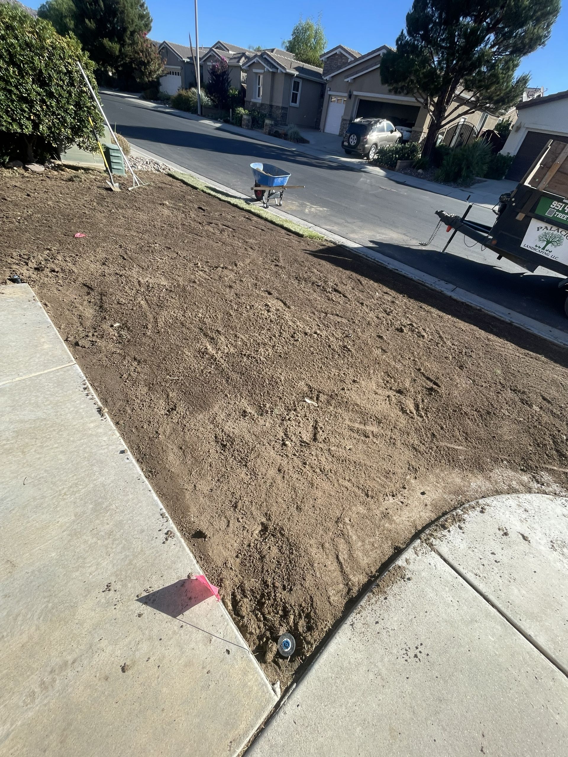 Dry dirt patch bordered by sidewalks beside a residential street, with houses and trees in the background.