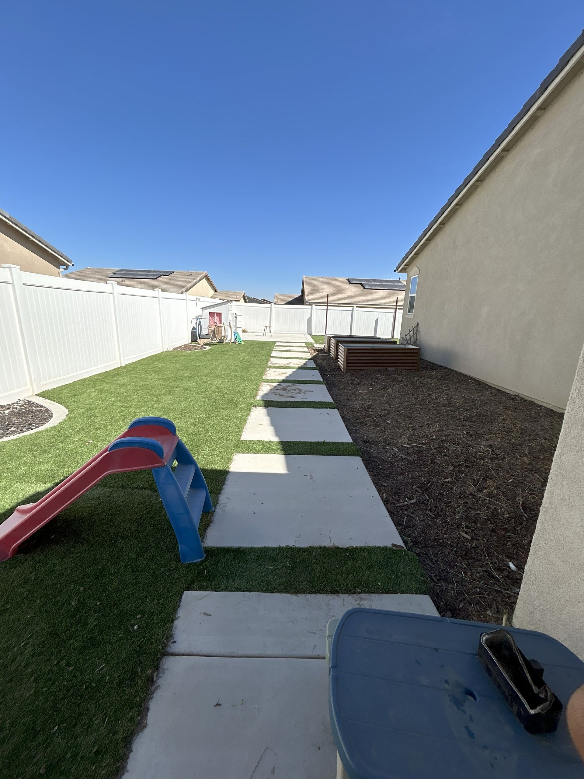 Narrow backyard with concrete stepping stones, artificial turf, white fence, beige wall, and a red-blue slide.