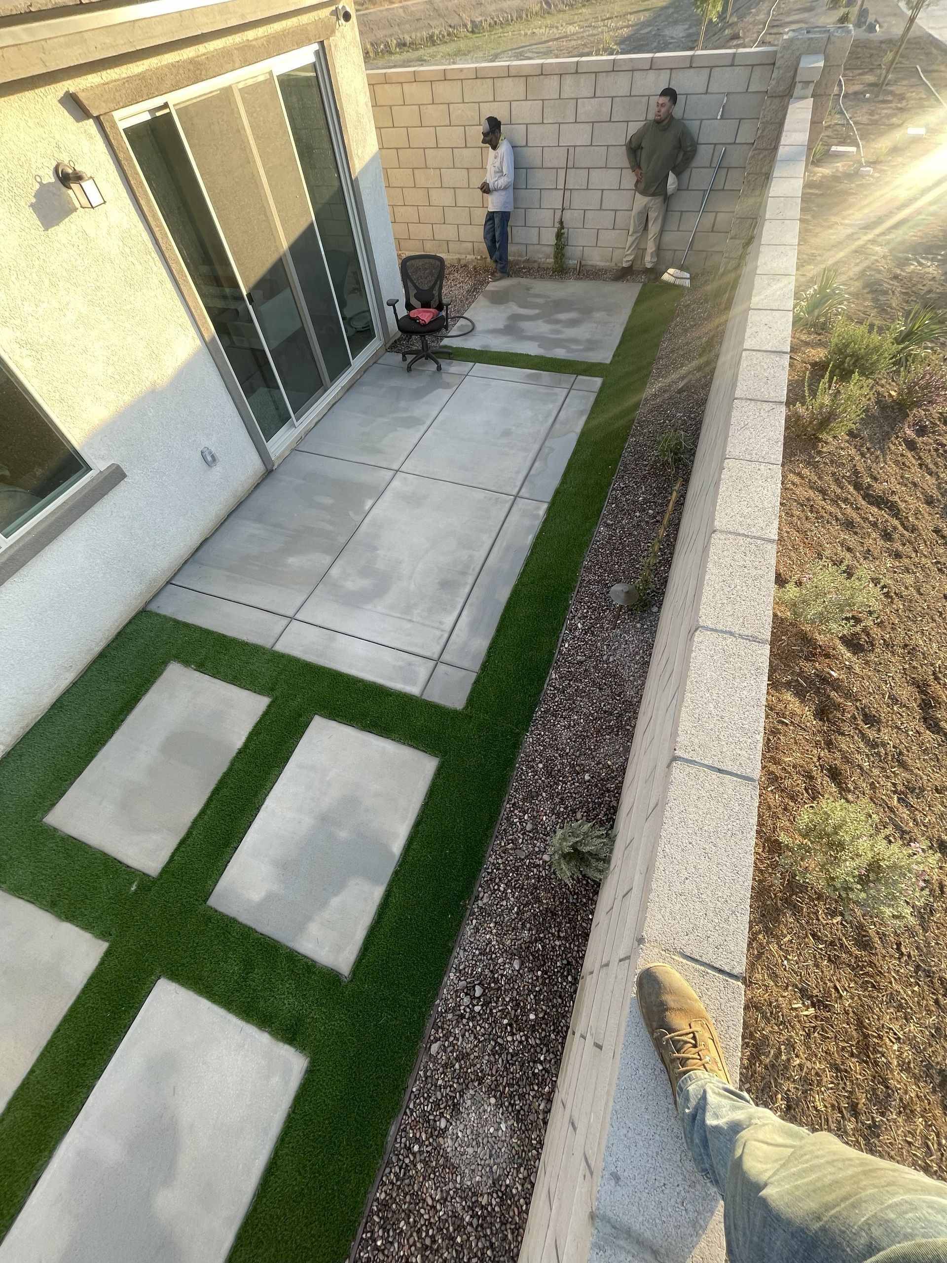 Narrow backyard with concrete stepping stones, artificial grass, stone walls, and two workers at the far end