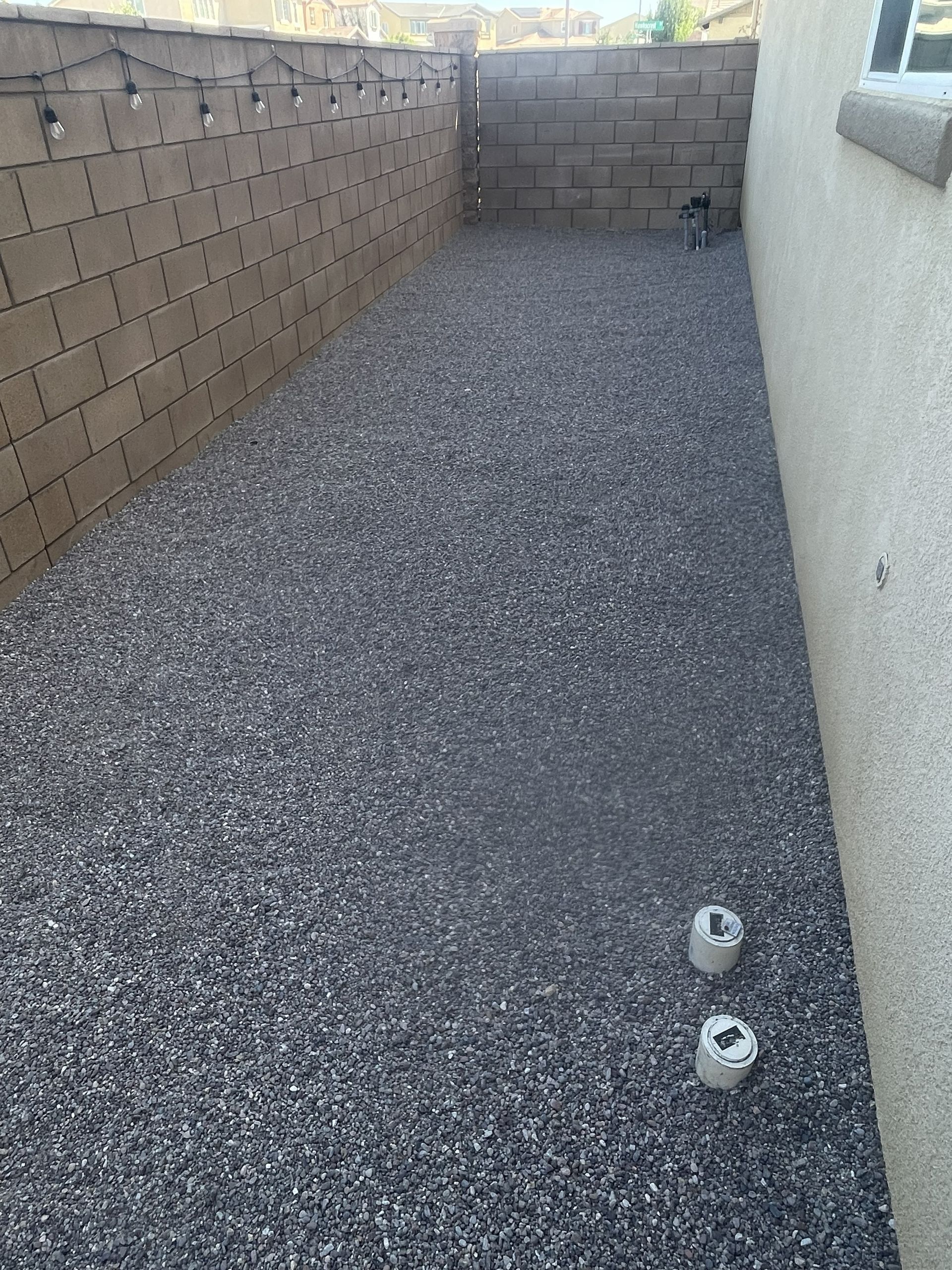 Narrow pea gravel walkway between a tan block wall and a white wall, with two small pipes near the foreground.