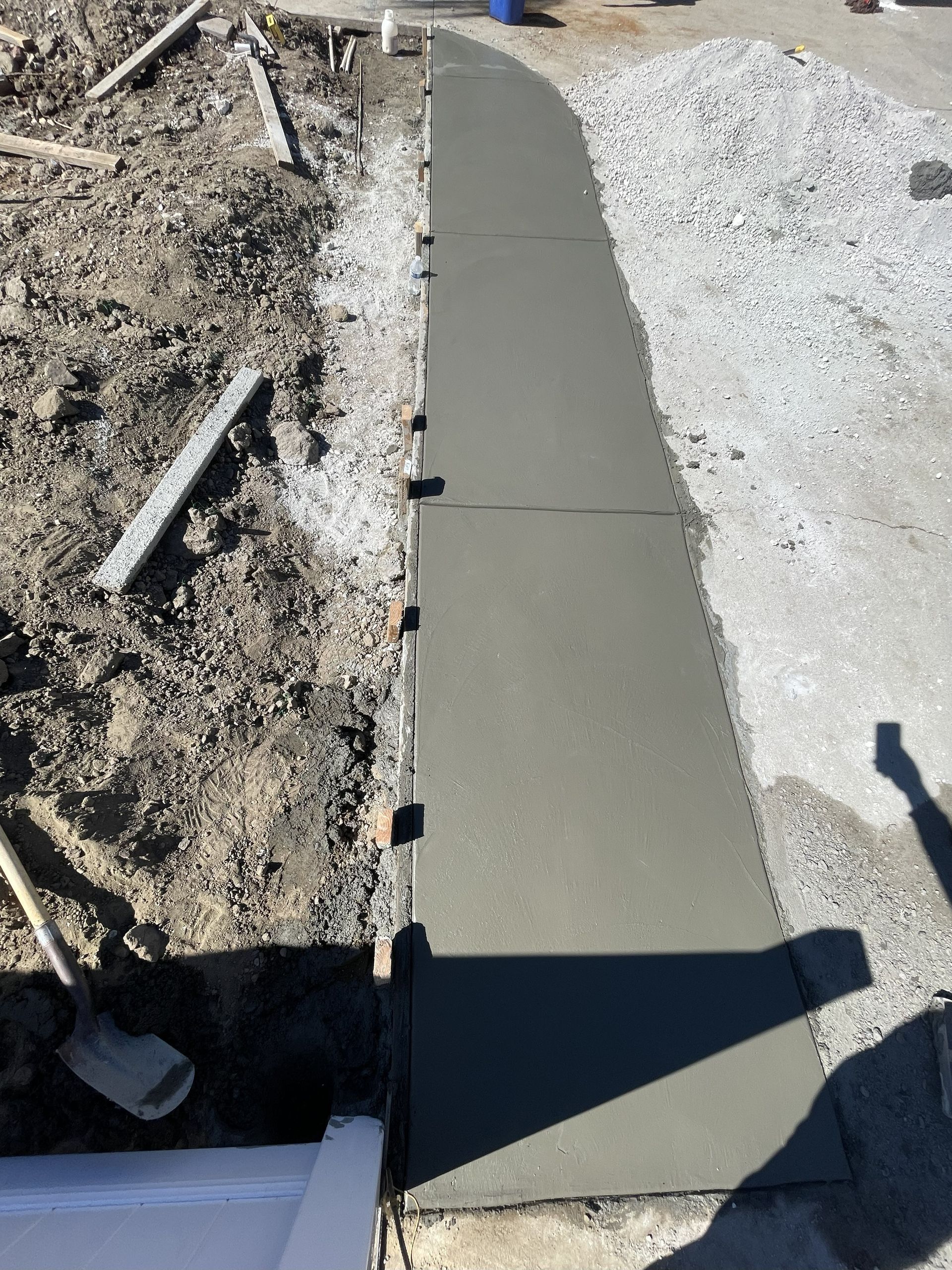 Fresh concrete walkway bordered by dirt and gravel, with an unfinished edge in sunlight