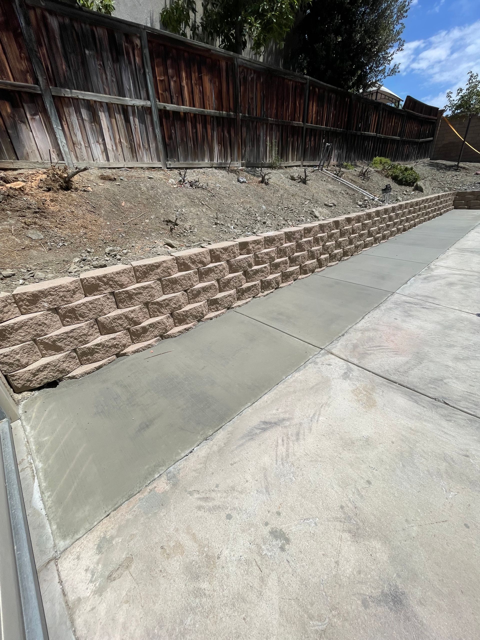 Concrete sidewalk beside a sloped retaining wall with wooden fence above and brick edging below