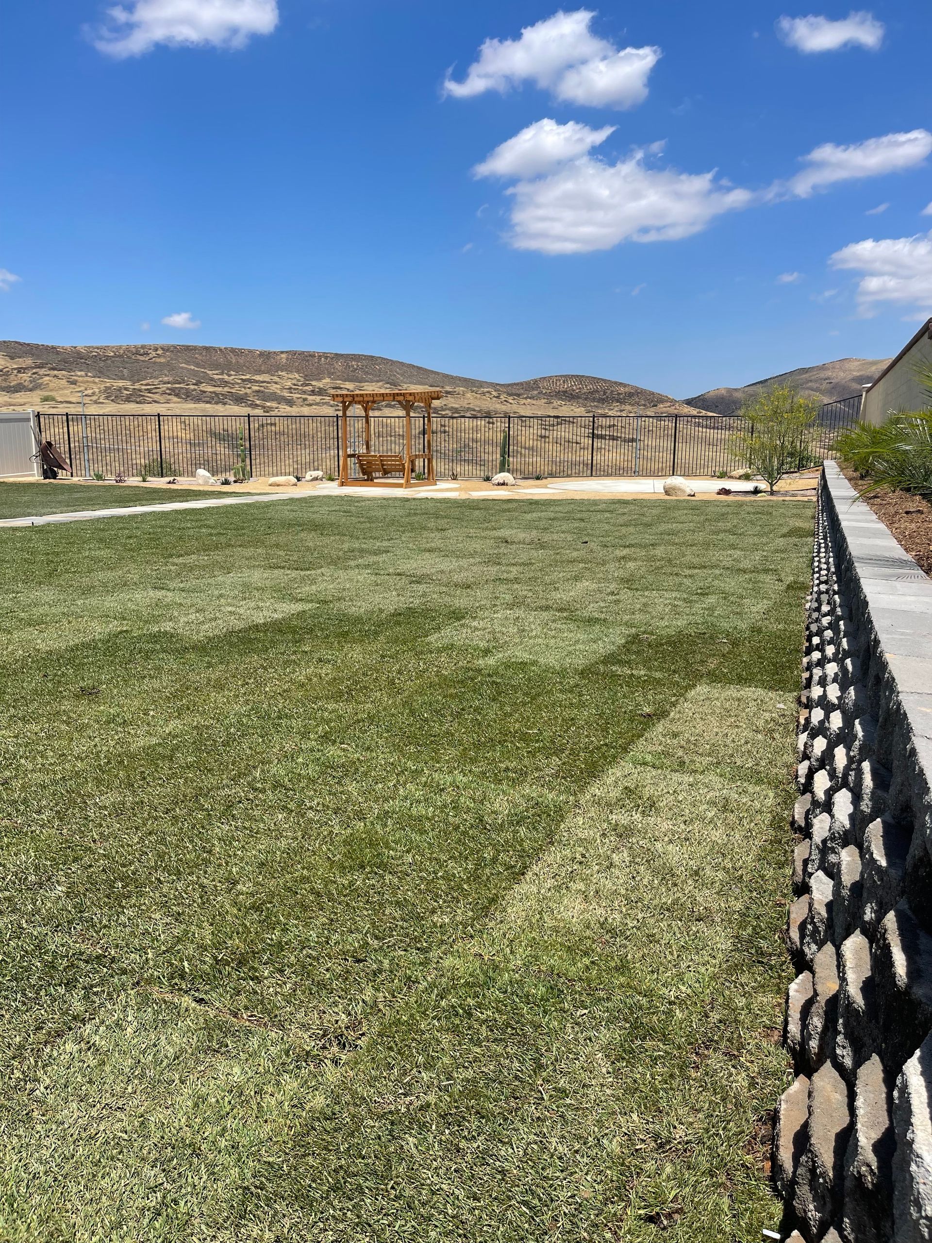 Stone wall and grassy courtyard under a blue sky with clouds, lined by a fence on the right