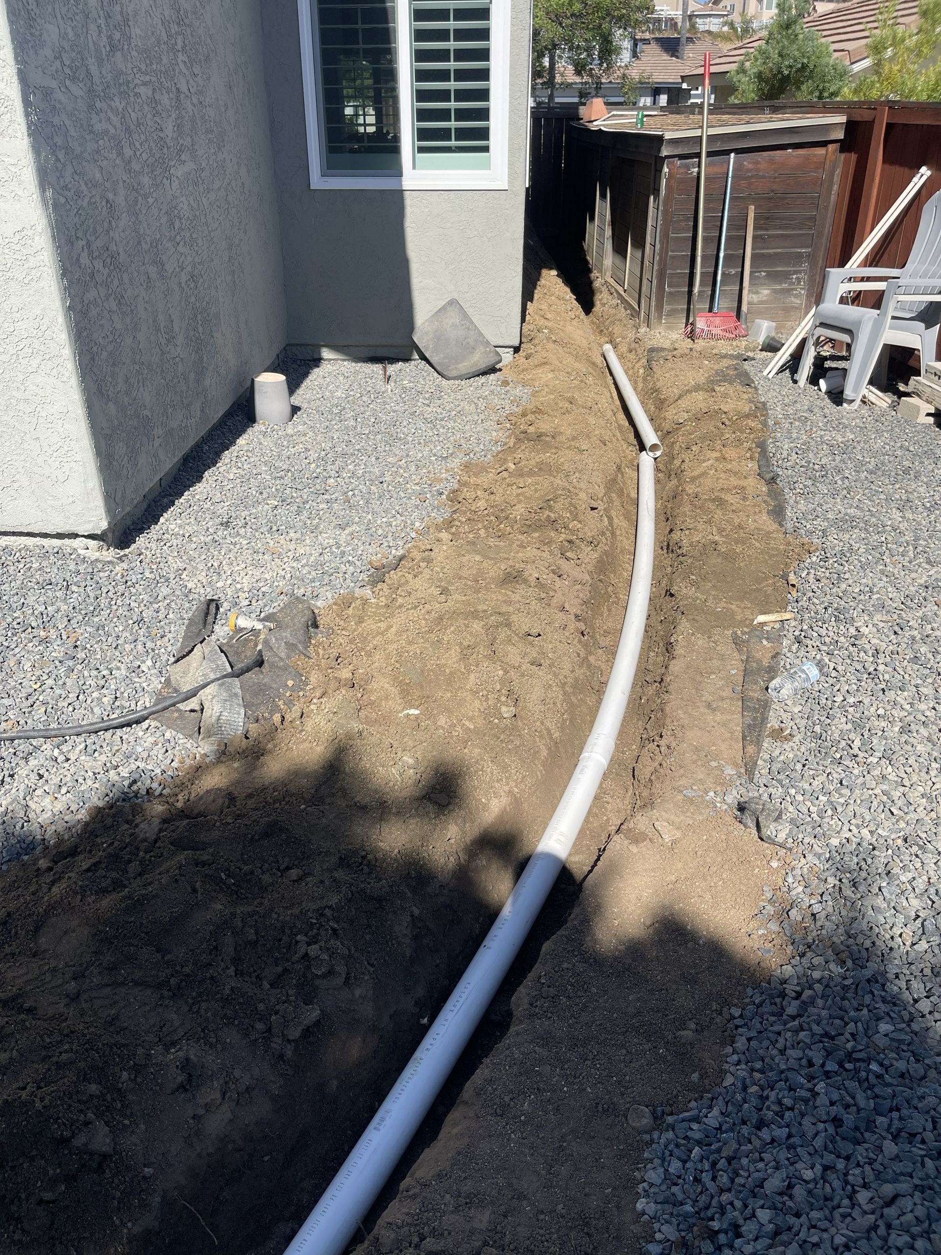 White drainage pipe laid along a dirt trench beside a house in a gravel yard