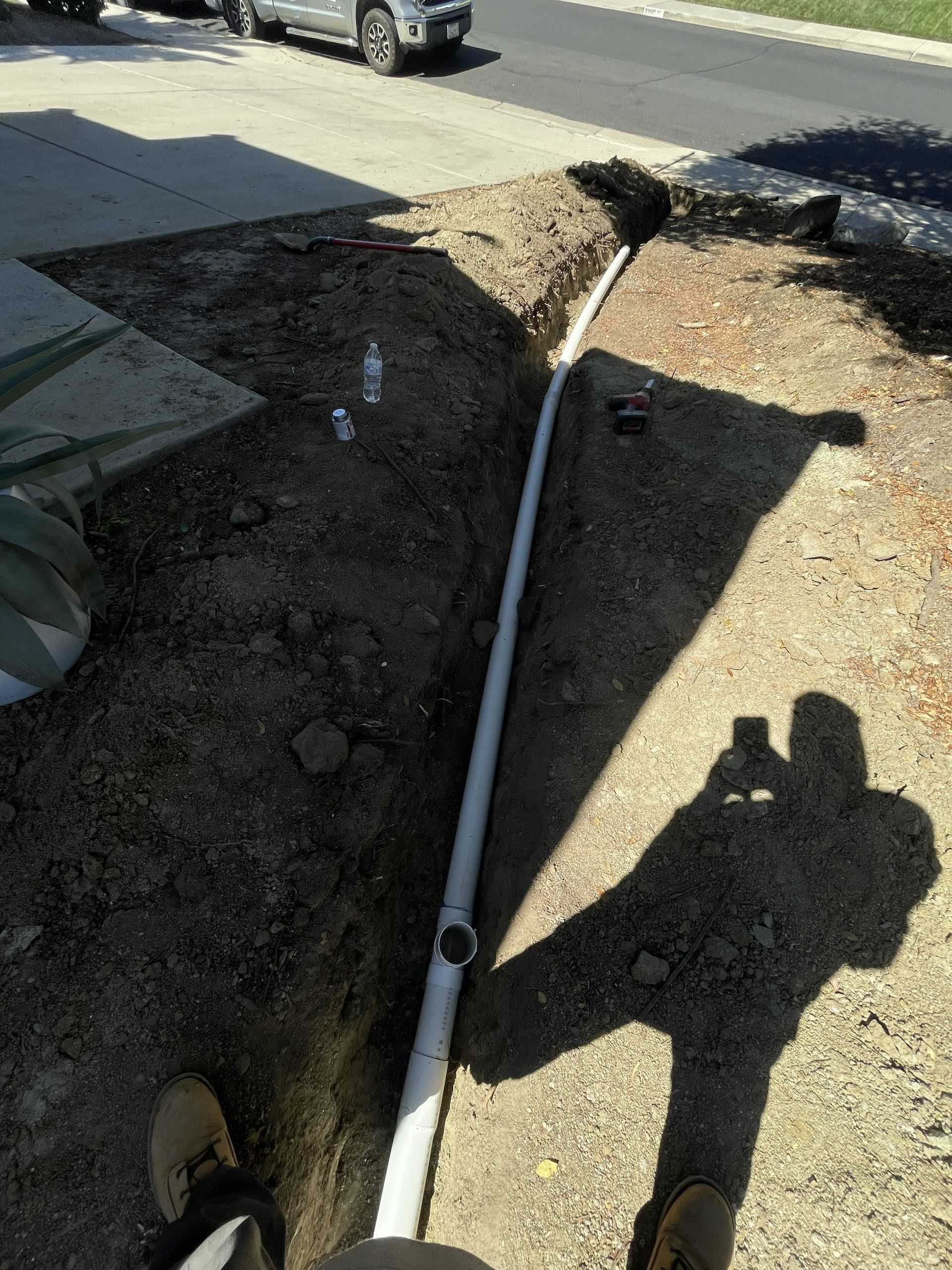 White pvc pipe lying across a dirt trench beside a sidewalk and parked cars.